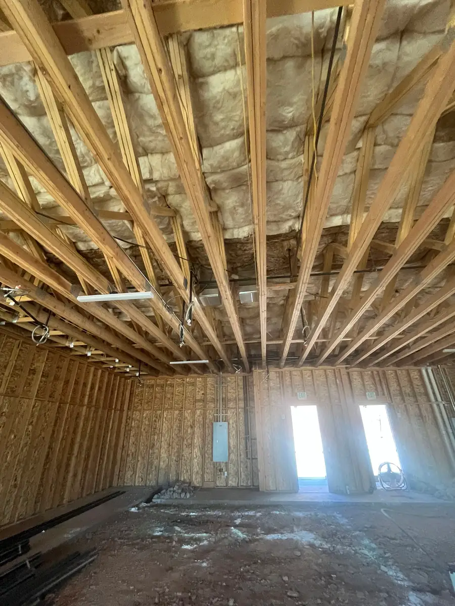 Upward view of wood roof trusses with insulation above them inside the Agave Animal Hospital building in St. George, Utah, with hanging light fixtures, bare stud walls, and two bright door openings below.