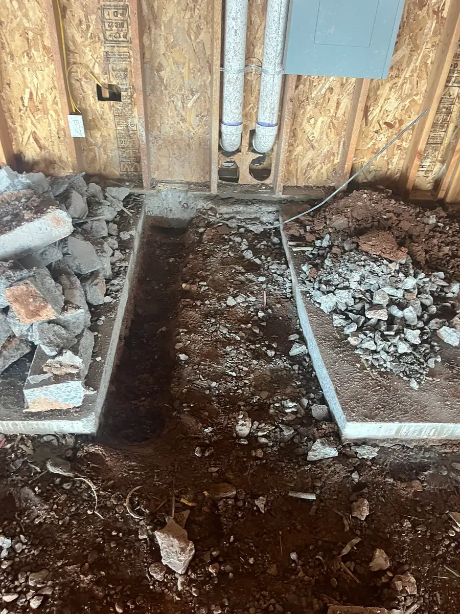 Close view of a trench cut through a concrete floor slab exposing soil and gravel beneath, with two white pipes running up inside a stud wall and broken-out concrete piled to one side.