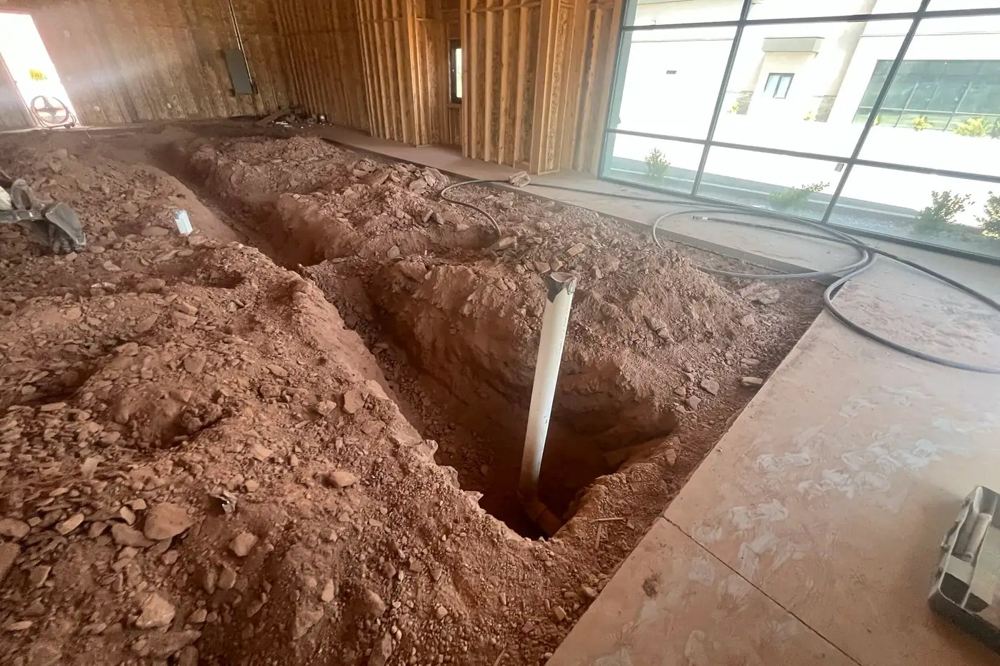 An open trench cut through red soil inside the Agave Animal Hospital building in St. George, with a white pipe standing up out of the trench, bare wood wall framing behind it, and a full-height glass storefront letting in daylight.