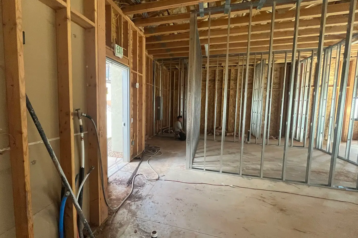 A wood-framed wall with blue and white tubing, a spigot with a hose attached, and a black pipe leaning against it, beside an open exterior doorway under an exit sign, with metal-stud rooms in the space beyond.