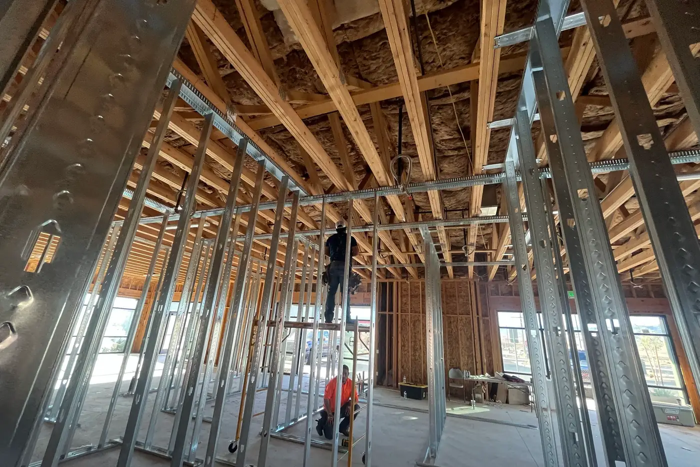 Two construction workers framing interior walls inside the Agave Animal Hospital building in St. George, Utah, one standing on a rolling scaffold and one kneeling at its base, surrounded by metal studs under wood roof trusses and insulation.