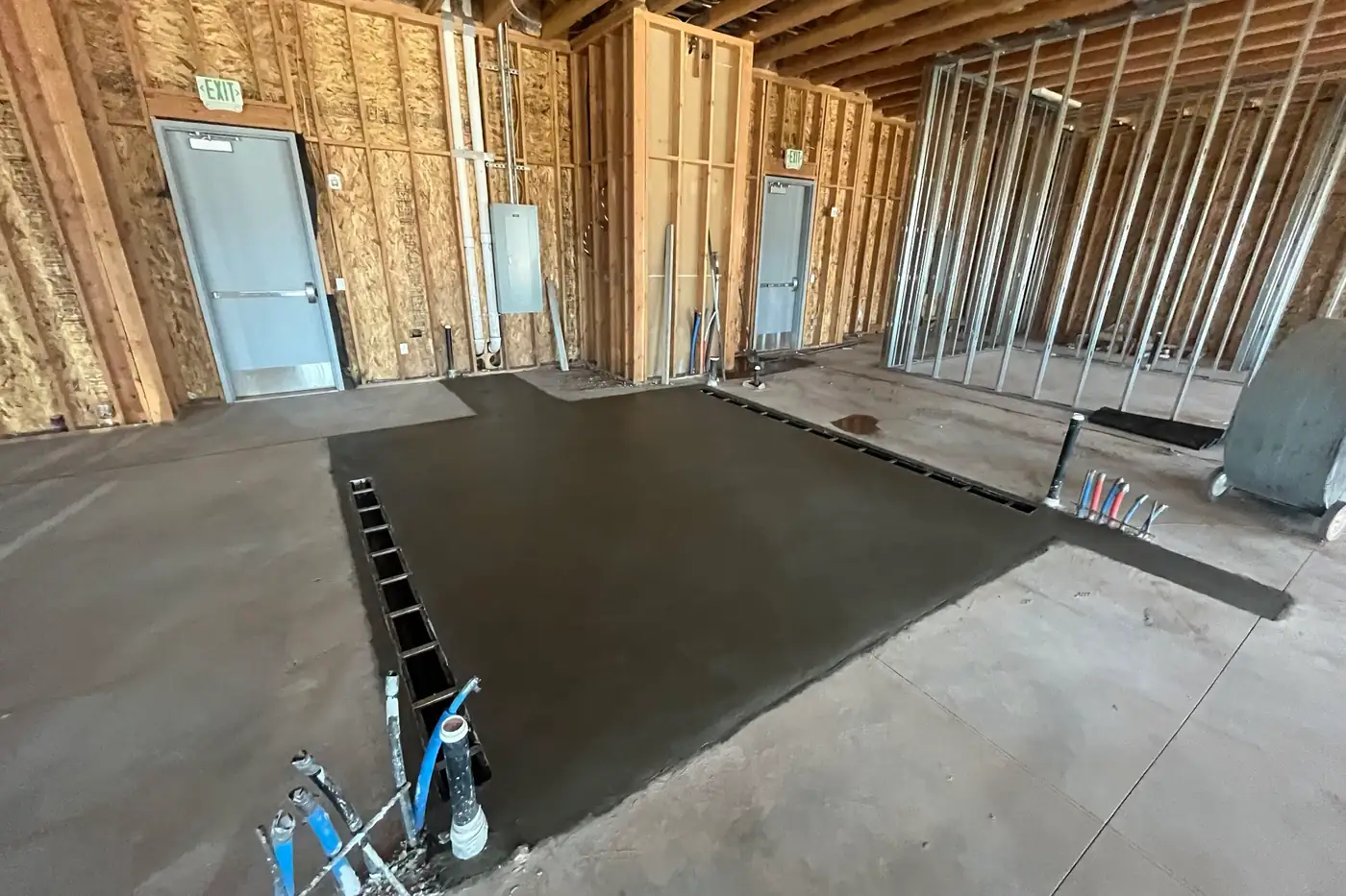 Another view of the darker freshly poured floor section with a slotted channel drain along its edge, two gray metal doors with exit signs on the wall behind, and metal-stud framing at right.