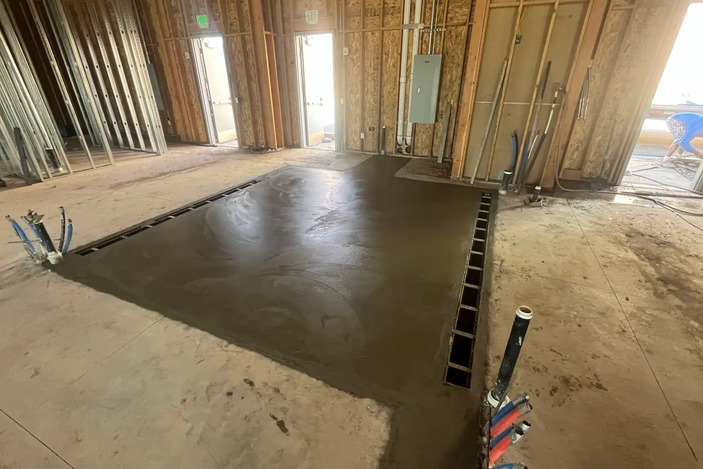 A rectangle of freshly poured, darker concrete set into the finished floor, with slotted channel drains along two edges, a gray panel box on the wall behind it, and open doorways letting in daylight.