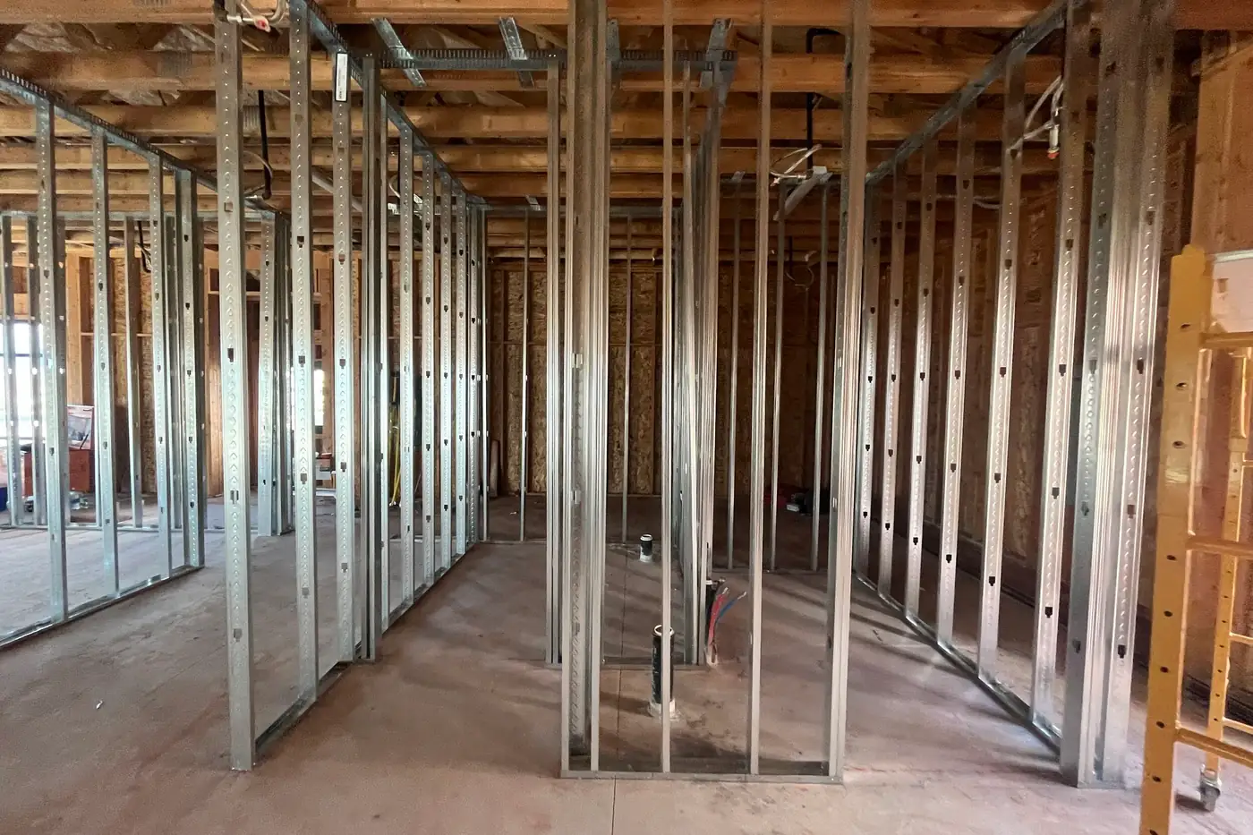 Looking through a series of metal-stud framed openings toward the back of the building, with a black pipe rising from the concrete floor and a yellow ladder at the right edge.