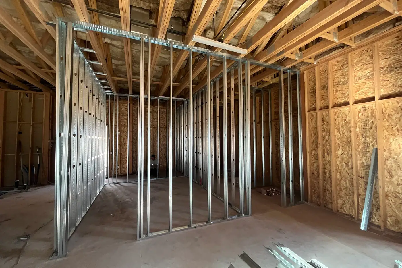 A rectangular room outlined by metal studs on a concrete floor, beneath exposed wood roof trusses and insulation, with sheathed wood walls behind it.