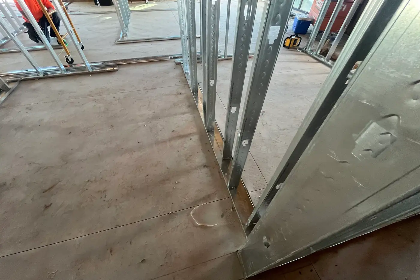 Low, close view of metal studs standing in a bottom track fastened to the concrete floor, with more wall framing and a rolling ladder in the background.