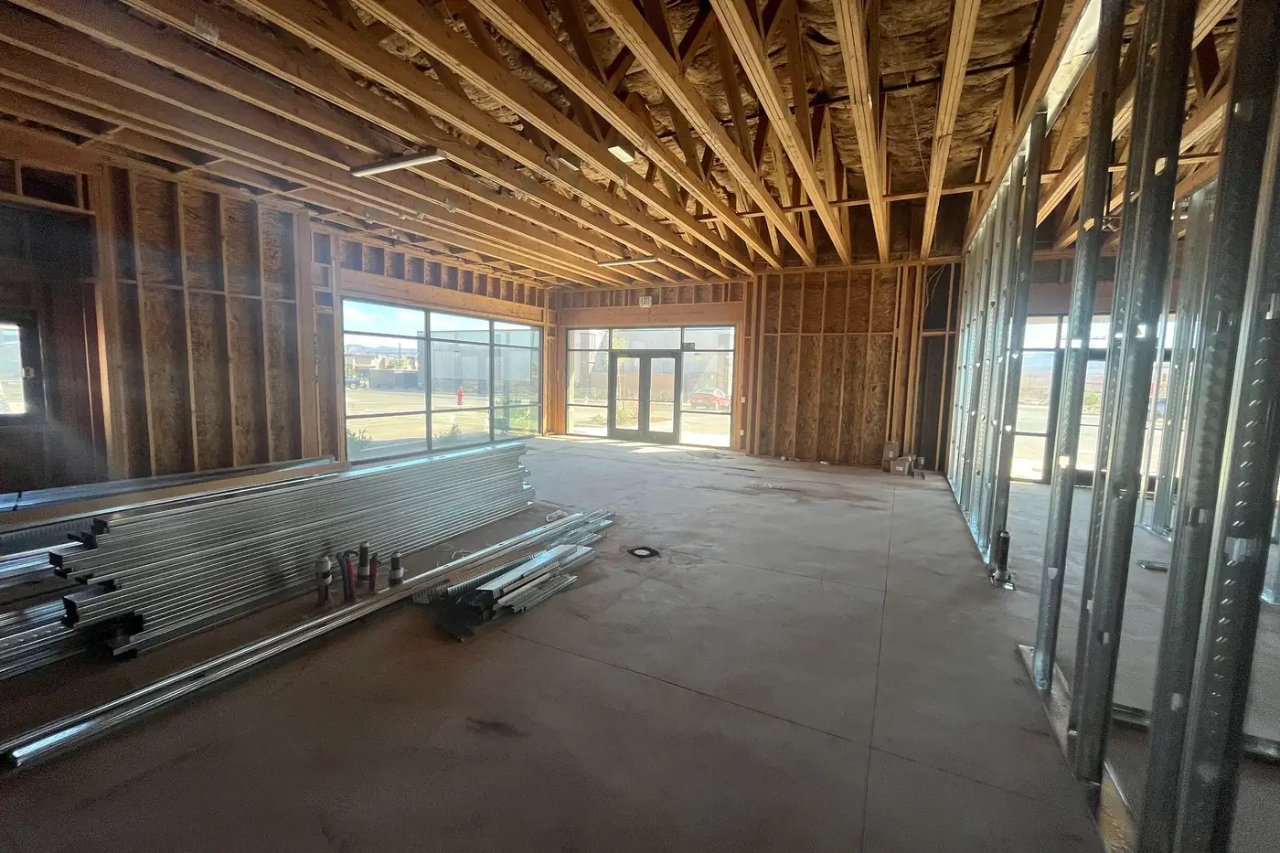 Wide view of a long front room inside the Agave Animal Hospital building, with bundles of metal studs on the concrete floor, storefront windows and glass double doors at the far end, and wood trusses overhead.