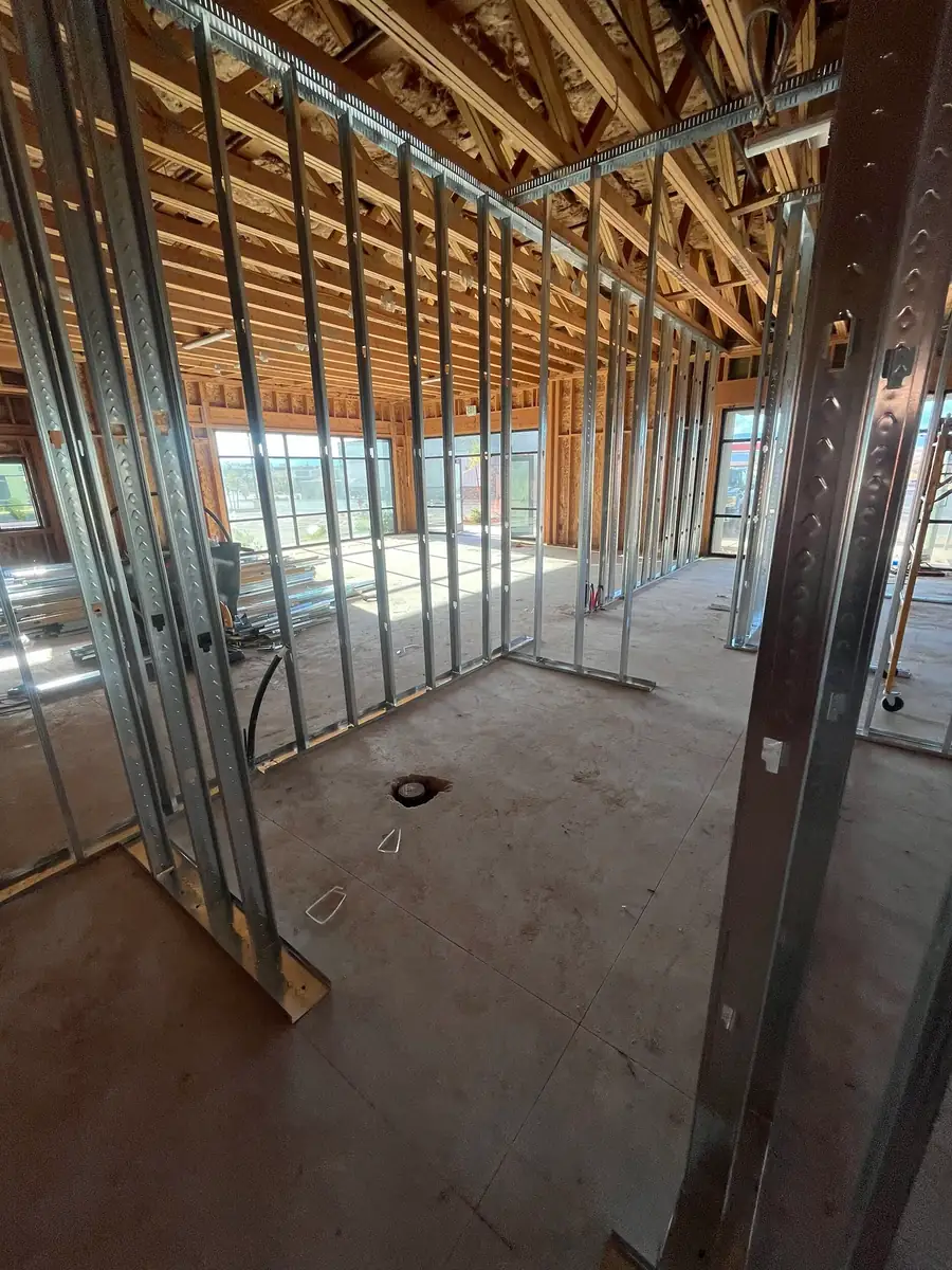 Portrait view from inside a room outlined in metal studs, with a round opening in the concrete floor and windows at the far end of the building.