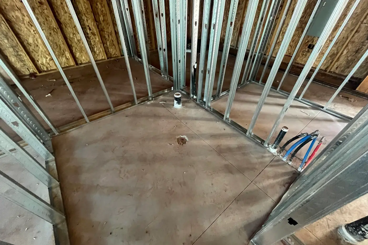 Blue and red tubing and a black pipe rising from the concrete floor at the base of a metal-stud wall, beside a round opening in the floor.