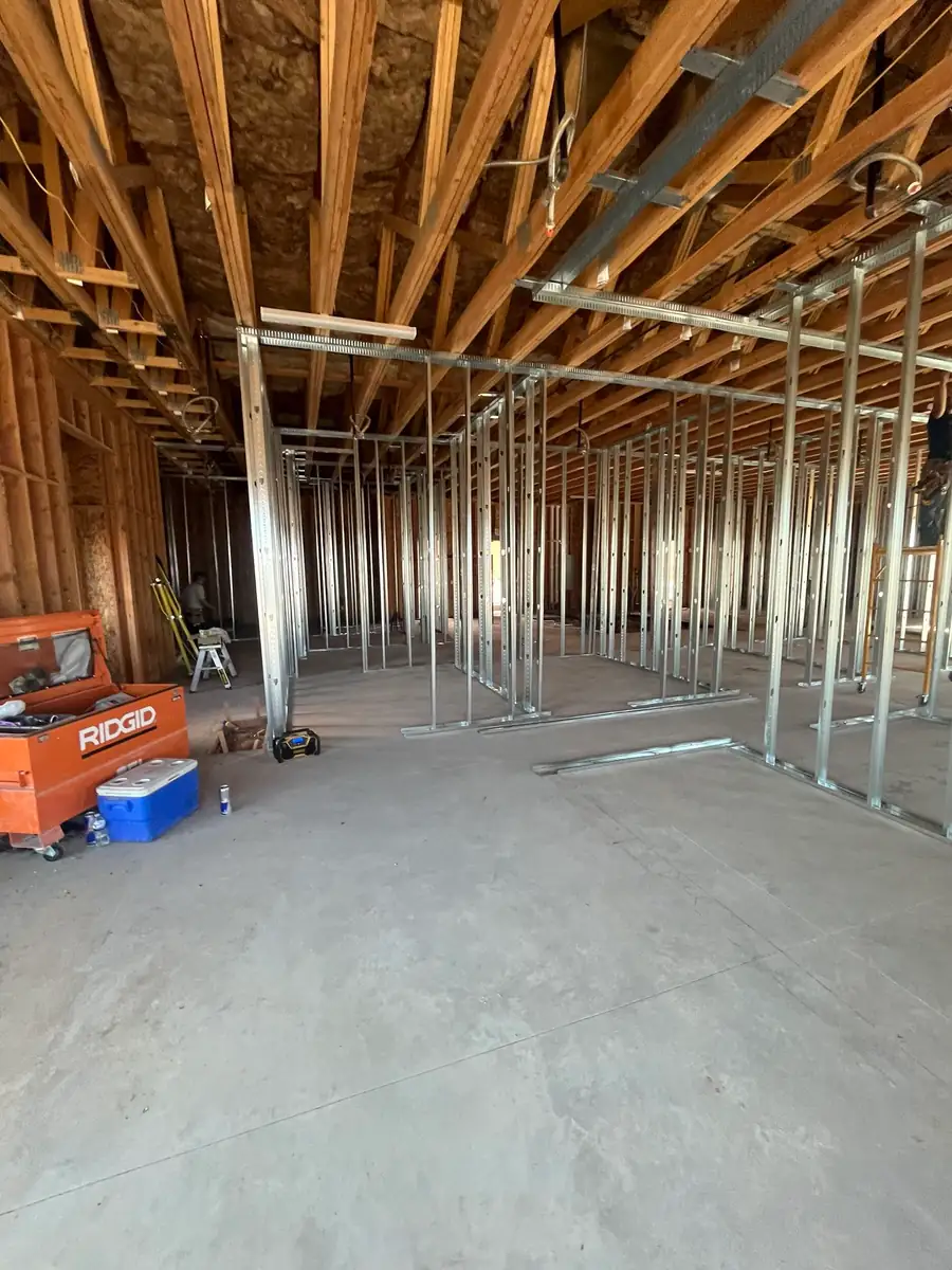 Portrait view down a long row of metal-stud walls under wood trusses, with an orange job box, a blue cooler and a stepladder at the left.