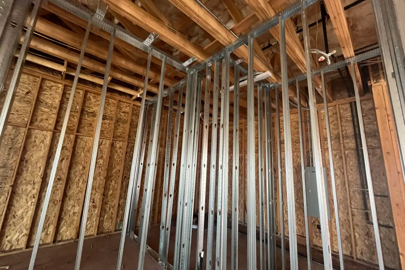 Upward view inside a room framed with metal studs, the tops of the studs meeting exposed wood trusses and insulation, with a gray panel box on the sheathed wall at right.