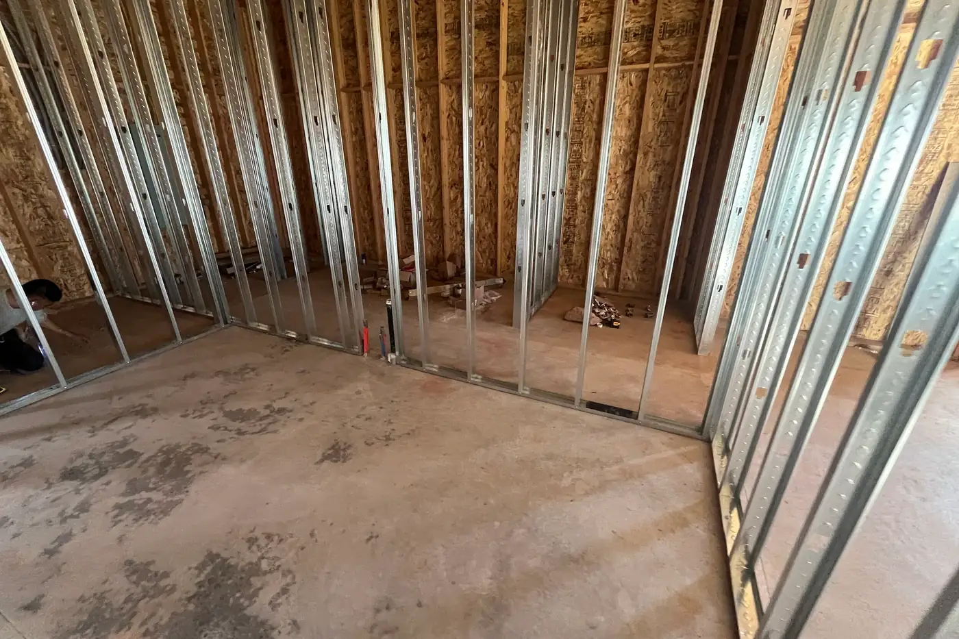 Inside a room outlined in metal studs against sheathed wood walls, with a worker crouched at the far left and building materials on the floor of the room beyond.