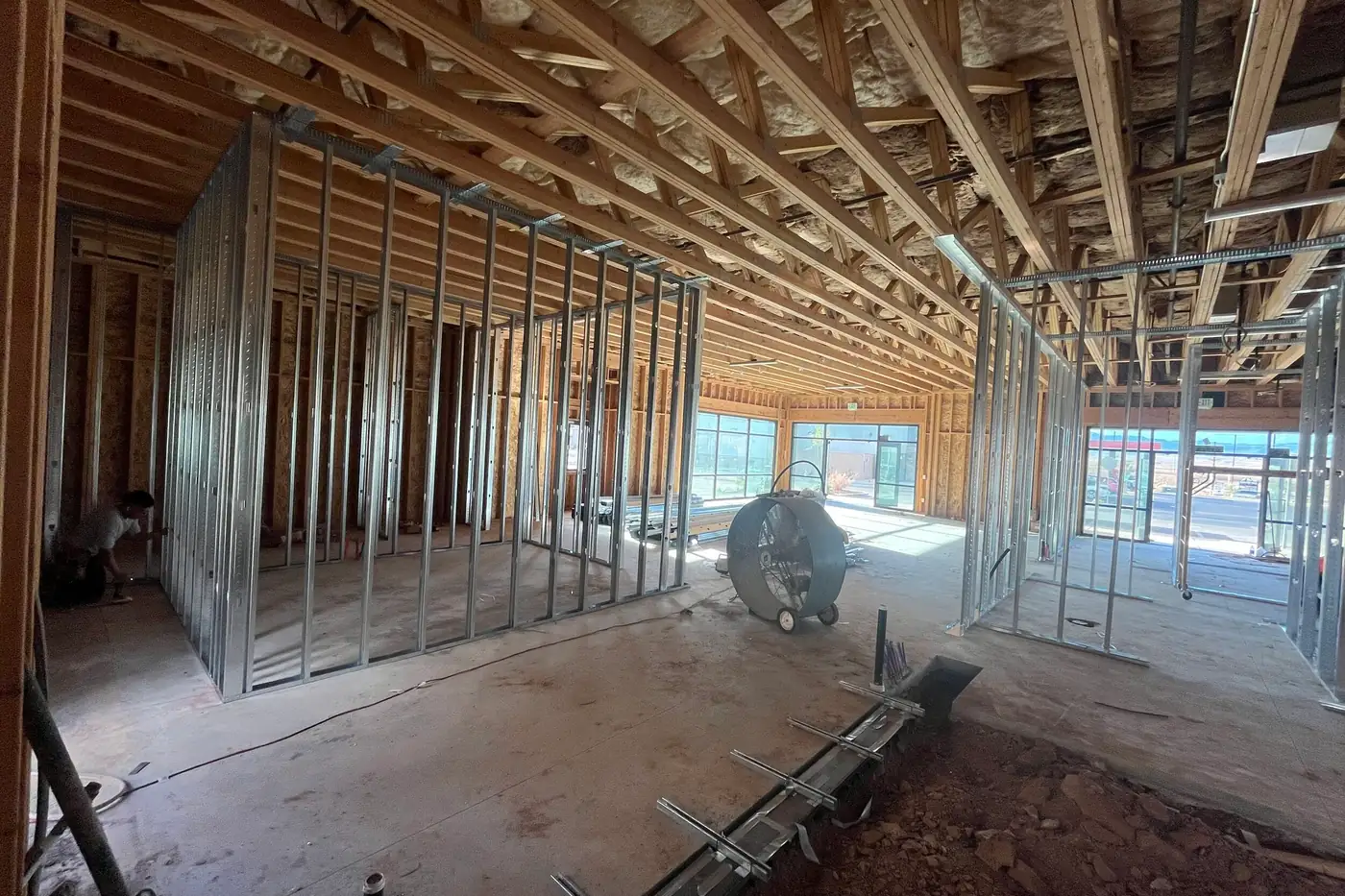 Wide interior view with metal-stud room framing on the left and right, a large drum fan in the middle, storefront windows at the far end, and a strip of open dirt and rock in the foreground with a long metal channel set along its edge.