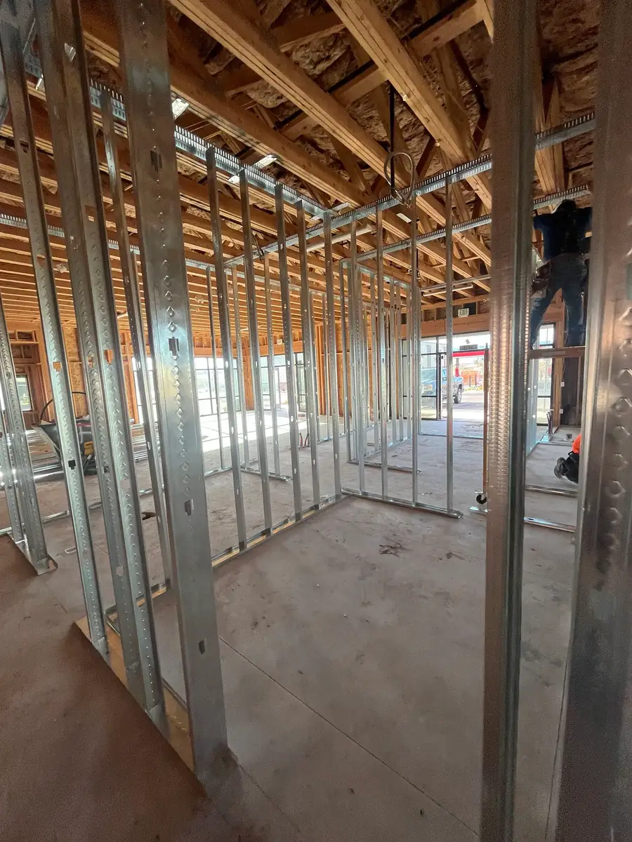 Portrait view of metal-stud room framing beneath wood roof trusses and insulation, looking toward glass doors at the far end of the building.