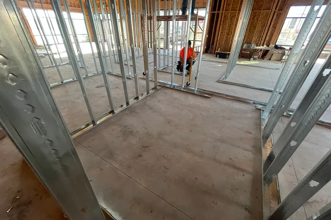 A worker in an orange shirt kneeling beside a rolling scaffold between metal-stud walls, with an empty framed room on a concrete floor in the foreground.
