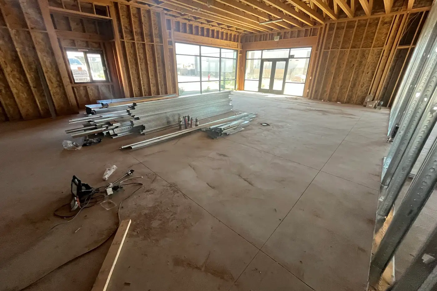 Bundles of metal studs and track lying on a concrete floor in a room with storefront windows, glass doors, and a work light and cords in the foreground.